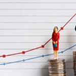 Miniature people toys standing on a pile of coins in front of a graph