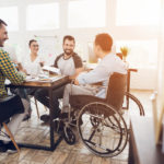 A man in a wheelchair communicates cheerfully with employees of the office during a business meeting.