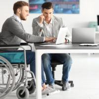 disabled businessman in wheelchair with coworker on laptops disabled businessman in wheelchair with coworker on laptops