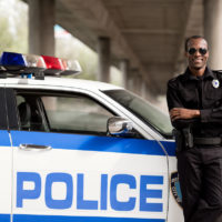 handsome african american police officer with crossed arms leaning back on car and looking at camera