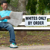 A non-white man sits on a bench in a park reserved for whites