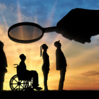 Employer's hand looks through the magnifying glass to an invalid in a wheelchair waiting for an interview for work