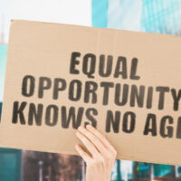 The phrase " Equal opportunity knows no age " on a banner in men's hand with blurred background. Equality on workplace. Office. Job. Employment. Work. Human rights. Difference. No discrimination