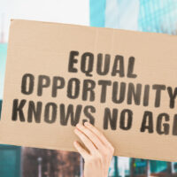 The phrase " Equal opportunity knows no age " on a banner in men's hand with blurred background. Equality on workplace. Office. Job. Employment. Work. Human rights. Difference. No discrimination The phrase " Equal opportunity knows no age " on a banner in men's hand with blurred background. Equality on workplace. Office. Job. Employment. Work. Human rights. Difference. No discrimination