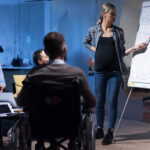 Handicapped young man with his colleagues working in office. Disabled businessman in the wheelchair and his colleagues listetning presentation of their pregnant female colleague.