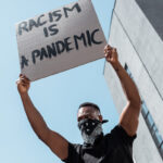 low angle view of african american man with scarf on face holding placard with racism is a pandemic lettering near building