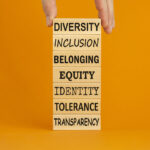 Diversity belonging inclusion equity identity tolerance transparency words written on wooden block. Male hand. Beautiful orange background. Diversity, inclusion and belonging concept.