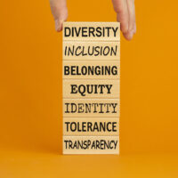 Diversity belonging inclusion equity identity tolerance transparency words written on wooden block. Male hand. Beautiful orange background. Diversity, inclusion and belonging concept.