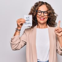 Middle age beautiful woman asking for equality holding paper with we are equal message smiling happy pointing with hand and finger to the side Middle age beautiful woman asking for equality holding paper with we are equal message smiling happy pointing with hand and finger to the side