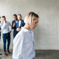 Male Colleagues Whispering Behind Back Of Unhappy Business Lady Indoors