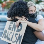 Young african woman hugging a caucasian woman after a protest - Northern woman with end racism bannner in her hands - Concept of no racism