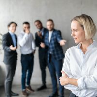 Male Coworkers Whispering Behind Unhappy Victimized Business Lady In Office