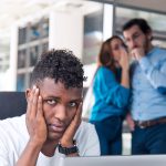 A Sad looking Black man is working on desk and there are two colleagues are gossiping at background behind of him