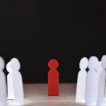 A single red paper cutout of a person standing centered and facing forward, flanked by two rows of similar white paper cutouts. The red figure stands out prominently against the white figures and a stark, solid black background.