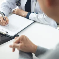 Doctor and patient at hospital office. Doctor working on diagnostic examination on men's health disease and writing on prescription record information document at medical clinic, close up Doctor and patient at hospital office. Doctor working on diagnostic examination on men's health disease and writing on prescription record information document at medical clinic, close up